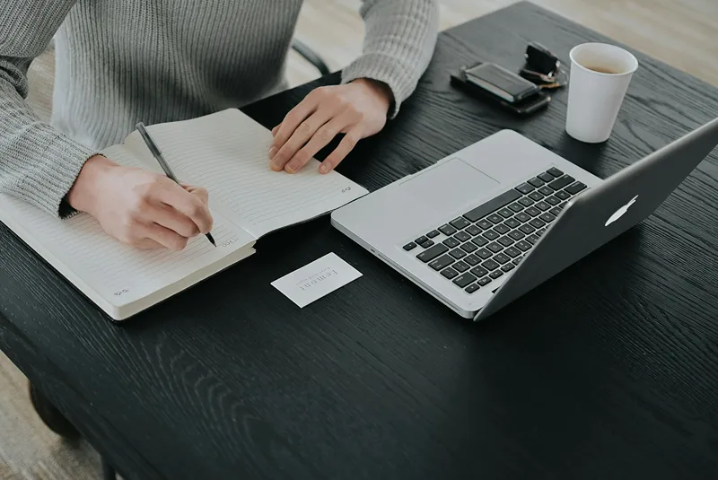Person taking notes in a notebook while working on a laptop, illustrating productive work habits and effective note-taking.